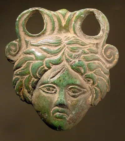 Henkelattasche eines Eimers "Beschlagstück in Form einer weiblichen Maske, wohl Gorgoneion, ... Oben Ansatz mit zwei großen Öffnungen und einer Volute jederseits. Hinten flach. Vollguß"