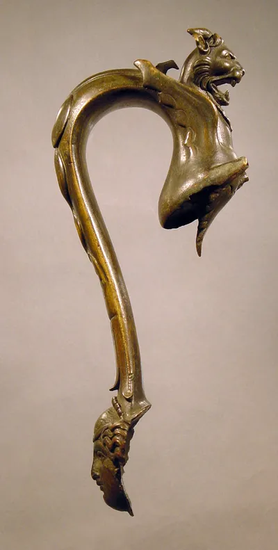 Griff eines Gefäßes. Oben verziert mit dem Kopf eines gehörnten und geflügelten Löwen, unten mit einem weiblichen Kopf von vorn