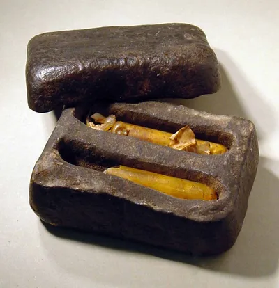 Behälter (aus einer weichen harzigen Masse), in dem zwei Glasröhrchen liegen, je in einer Bettung; die Glasröhrchen sind oben und unten verschlossen und enthalten eine gelbe Flüssigkeit.