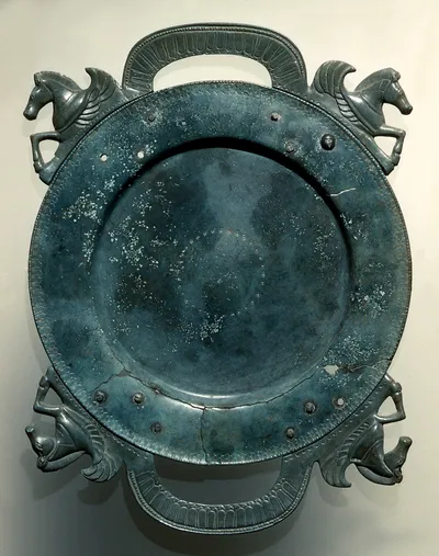 Teller (Teller mit bogenförmigen Henkeln, Flacher Teller mit zwei bogenförmigen, mit Nieten befestigten Griffen, an den Enden die Vorderteile von Flügelpferden, glänzende grüne Patina, Rand gebrochen)