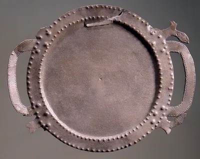 Runder Teller aus flachem Blech mit zwei Henkeln in Schleifenform; der Rand mit kleinen Buckeln verziert