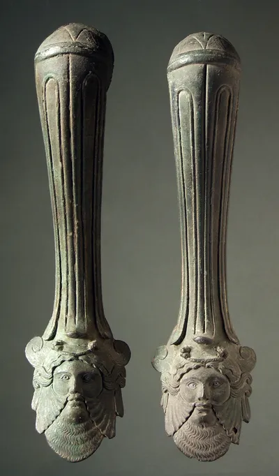 Zwei Gefäßhenkel (von einer römischen Amphora) mit bärtiger Maske, am Griffteil des Henkels drei schmale Längsblätter